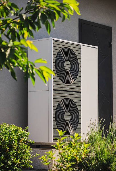 Modular heat pump unit for home heating and cooling installed next to a residential wall, surrounded by decorative plants and garden landscaping. High quality photo